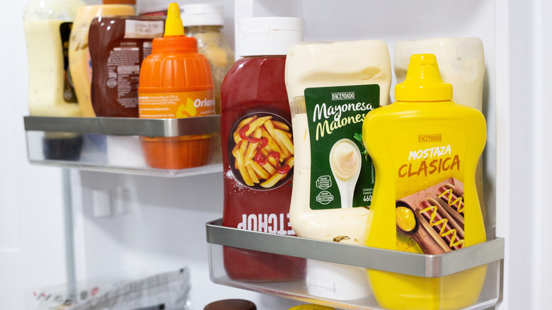 Bottles of ketchup, mayonnaise, and mustard sit inside the door of a refrigerator.