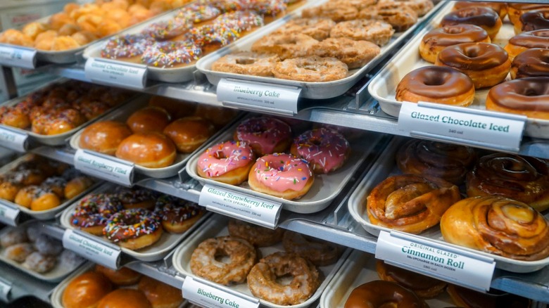 Donuts on display in Krispy Kreme store including chocolate iced, strawberry iced, blueberry cake, and cinnamon roll varieties