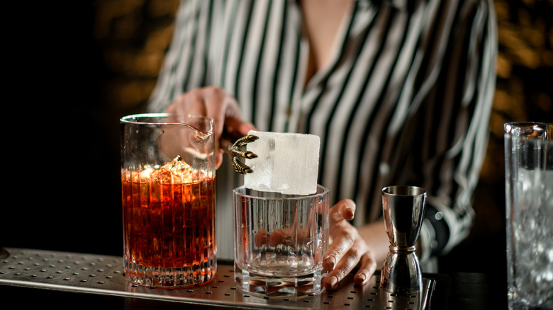 A close-up view of a female bartender holding large ice cube over glass. Mixing cup with drink standing nearby.