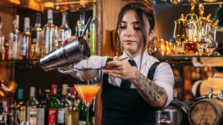 A young female bartender double-straining a cocktail behind the bar.