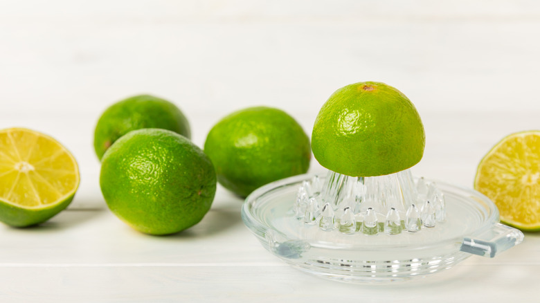 Whole limes on a white surface alongside a glass juicer with a halved lime on top.
