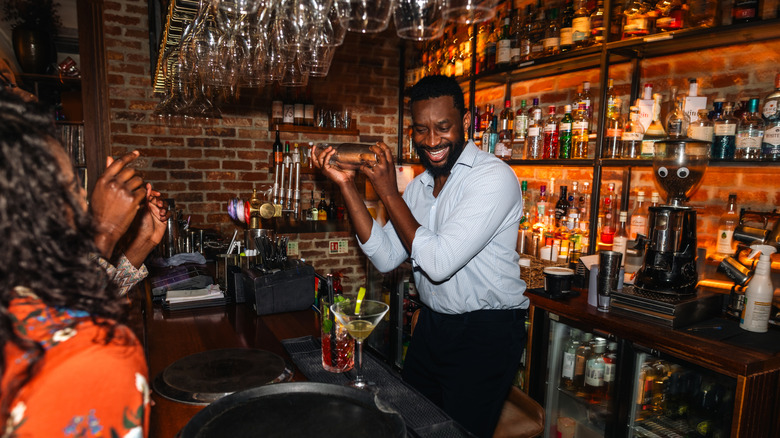 A wide over the shoulder shot of a smiling bartender energetically shaking a cocktail behind a lively brick-bar as clients cheer, creating a fun, festive atmosphere of nightlife.