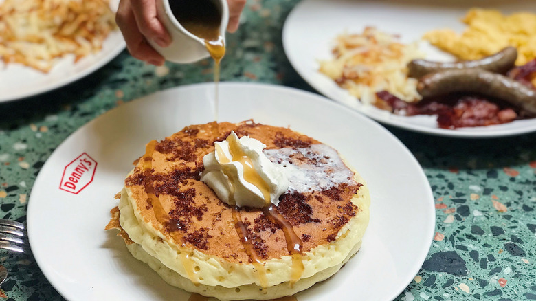 Denny's pancakes on a plate with someone pouring maple syrup on top.