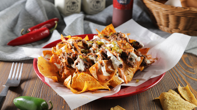 Brisket nachos with loaded toppings in a paper-lined basket with jalapenos, tortilla chips and other items surrounding it on a table.