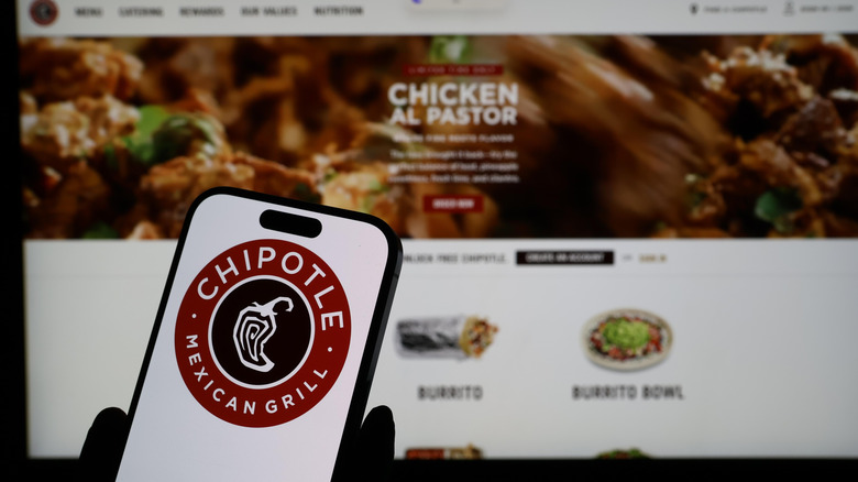 Someone holding a phone and accessing the Chipotle app with the company website on a computer screen in the background