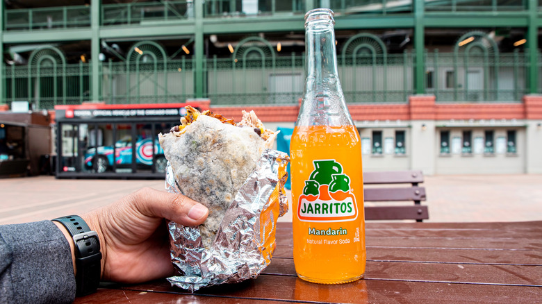 a hand holding a large, foil-wrapped burrito next to a bottle of Mandarin Jarritos with a building in the background