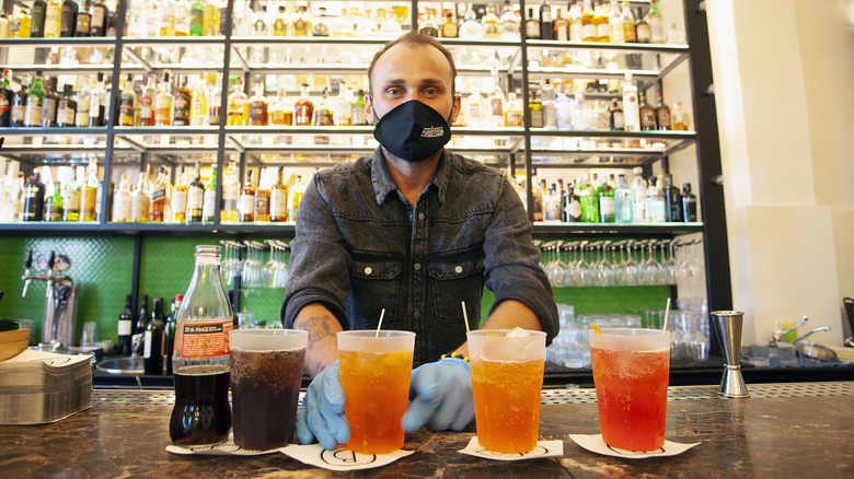 Bartender in mask making takeaway cocktails behind bar wall lined with bottles