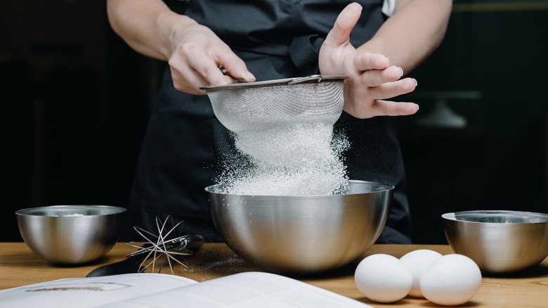 Hands sifting flour into a metal bowl next to eggs and bowls