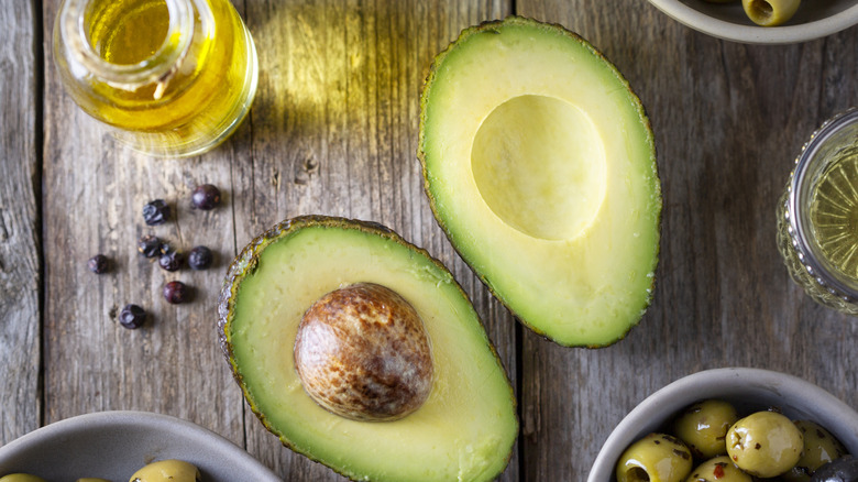 A split avocado on a wooden table showing the pit in one half with some olives and oil around the edges of the image