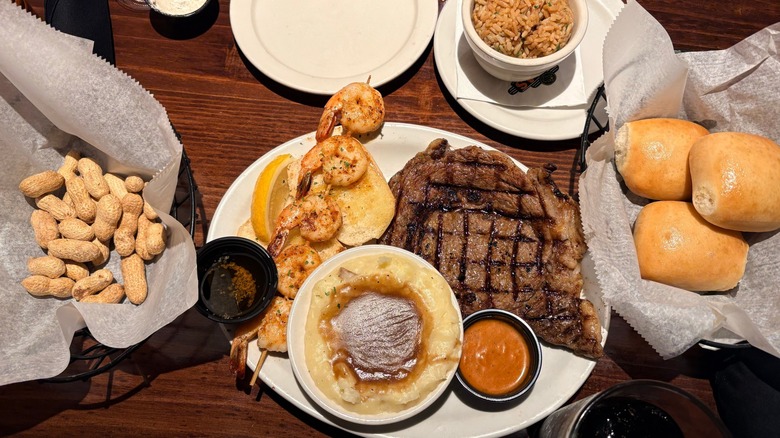 Top-view of a meal at Texas Roadhouse, including dinner rolls in a basket, skewered shrimp, basket of peanuts, mash potatoes and gravy, rice, and grilled meat
