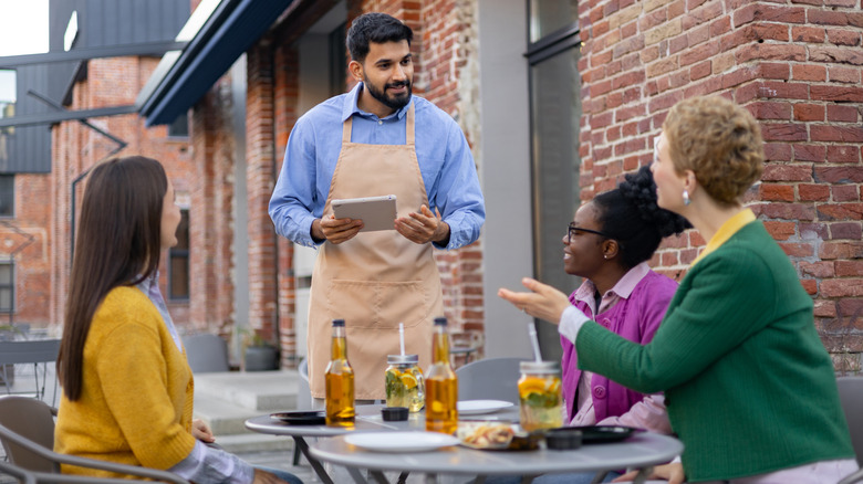 Three customers sitting at outdoor table at restaurant speaking to server with brick building in background