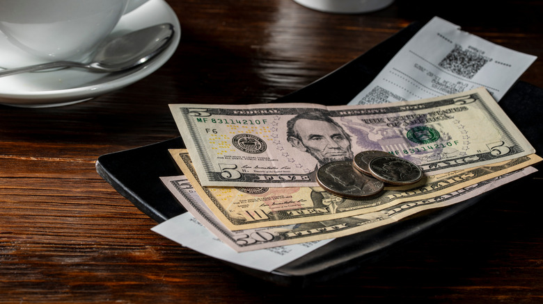 American dollar bills and coins piled on top of white restaurant receipt sitting on black tray.