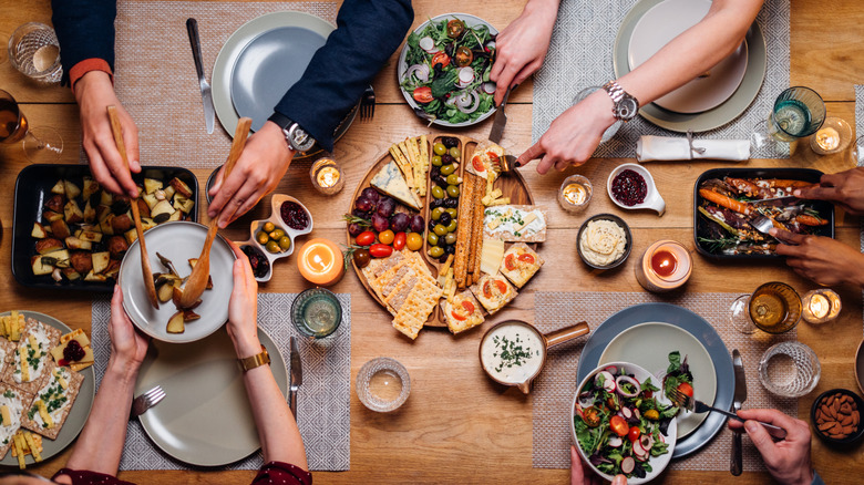 Four people sitting at set table sharing various food dishes including cheese platter, salads, and roasted vegetables