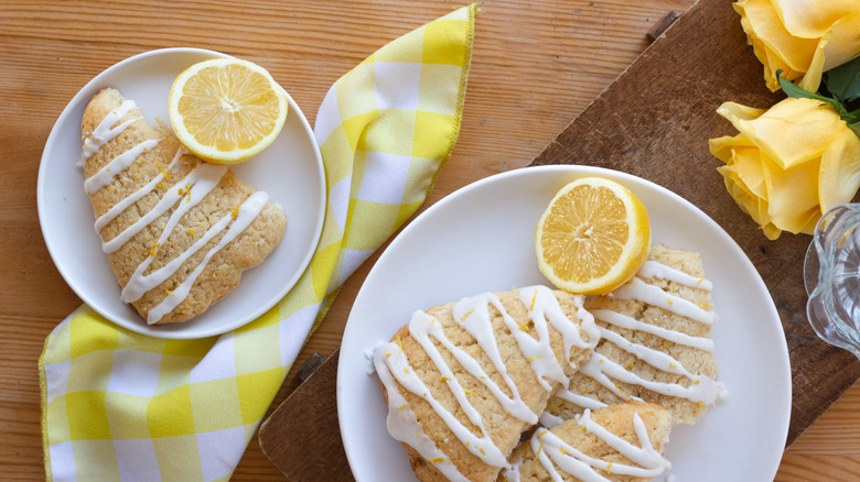 Scones with lemon zest and icing with halved lemons, yellow roses, and a yellow checkered napkin on a wooden table.