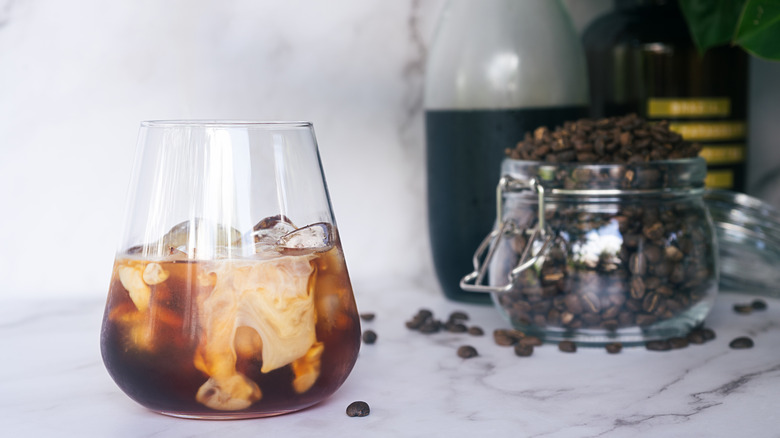 a glass of homemade cold brew coffee with an open jar of coffee beans in the background alongside a pitcher of cold brew