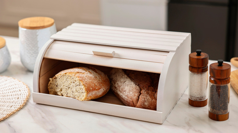 Seeded loaves in a white bread box on counter next to salt and pepper grinders