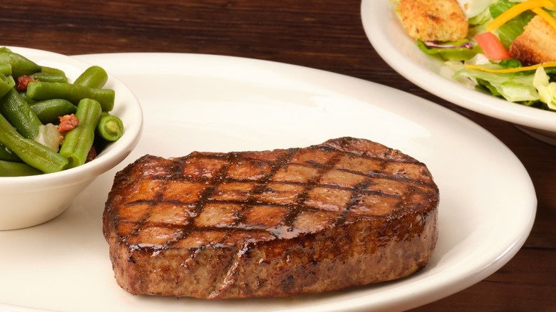 A Texas Roadhouse sirloin steak on a white plate next to a bowl of green beans and a side salad