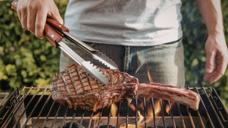 A person flipping a tomahawk steak on a grill with a pair of tongs