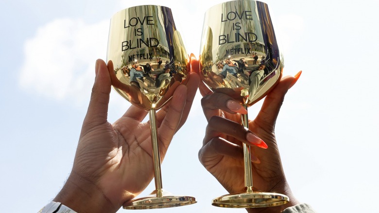 Two hands holding "Love Is Blind" branded gold wine glasses