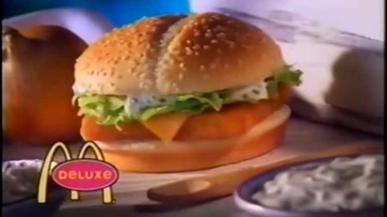 A McDonald's Fish Filet Deluxe sits on a wooden cutting board with an onion and tarter sauce