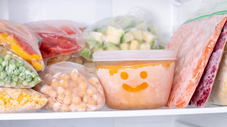 Several different kinds of frozen food arranged on the shelf of a home freezer, with a smiley-face drawn in the frost of a plastic tub