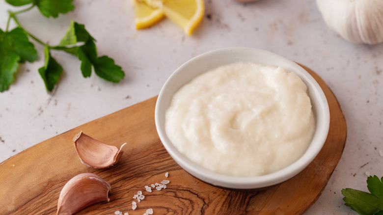 white bowl with toum in it with garlic cloves and salt on a wood board