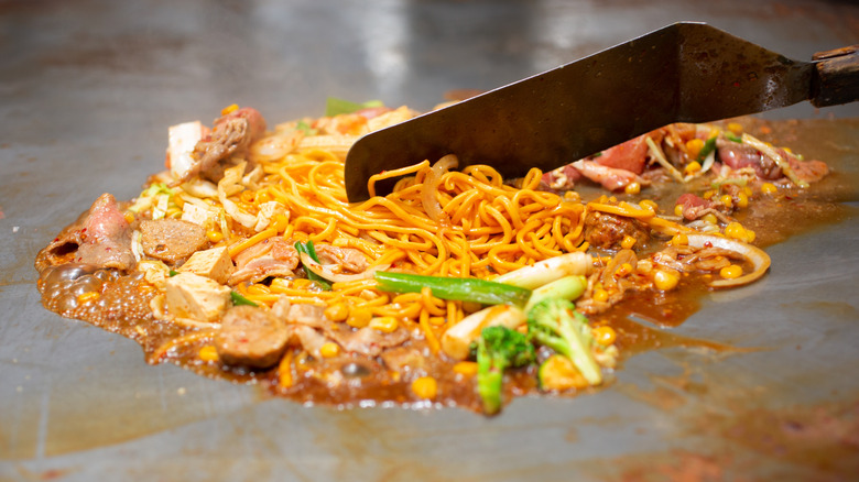 Noodle stir fry dish with meat and green vegetables cooking on flat top grill with metal spatula