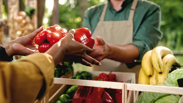 Someone handing two red bell peppers to another person at an outdoor market.