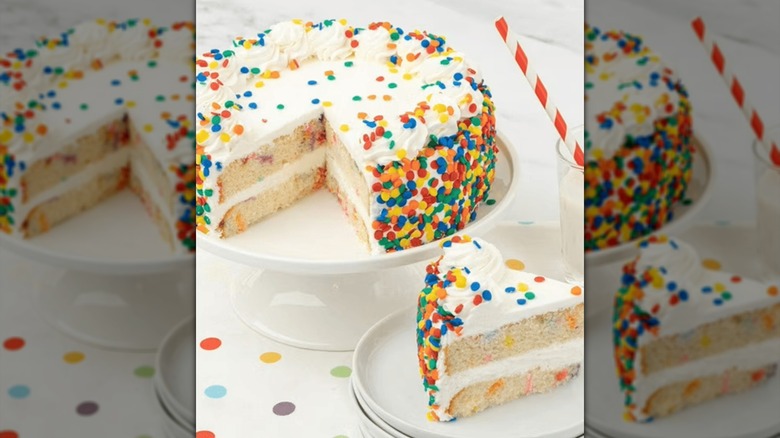 A round vanilla cake with a slice missing. Colorful confetti decorations are on top and on the sides.