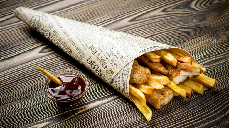 A fake newspaper shaped into a cone filled with fried fish and chips next to a small bowl of dipping sauce