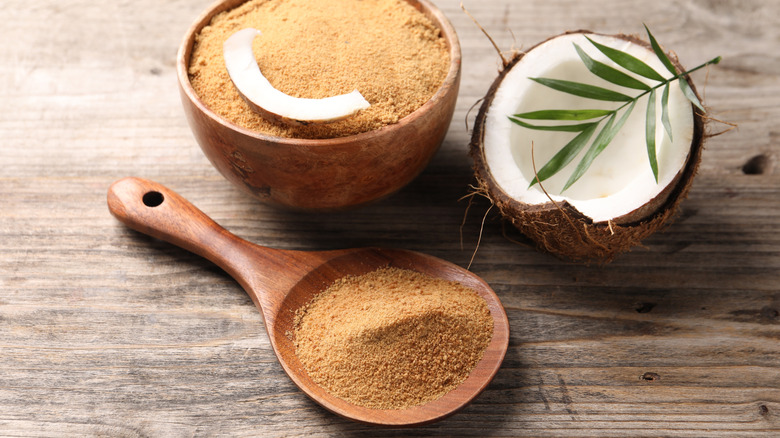 Coconut sugar in bowl on a wooden table and some in a spoon alongside a coconut half and palm foliage