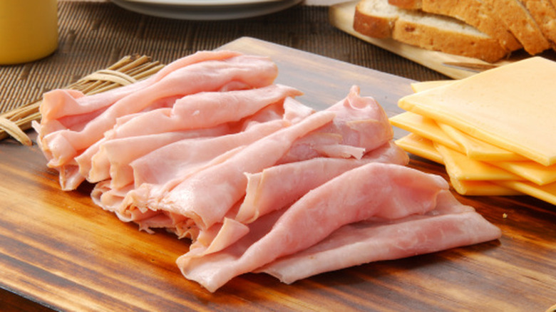 sliced deli ham on a wooden cutting board with slices of cheese and bread behind it