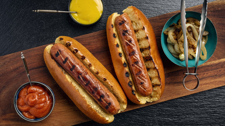 grilled hot dogs and buns on a wooden board with toppings alongside