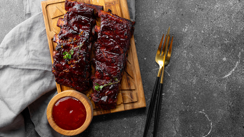 Ribs on a wooden board next to a bowl of sauce and a knife and fork