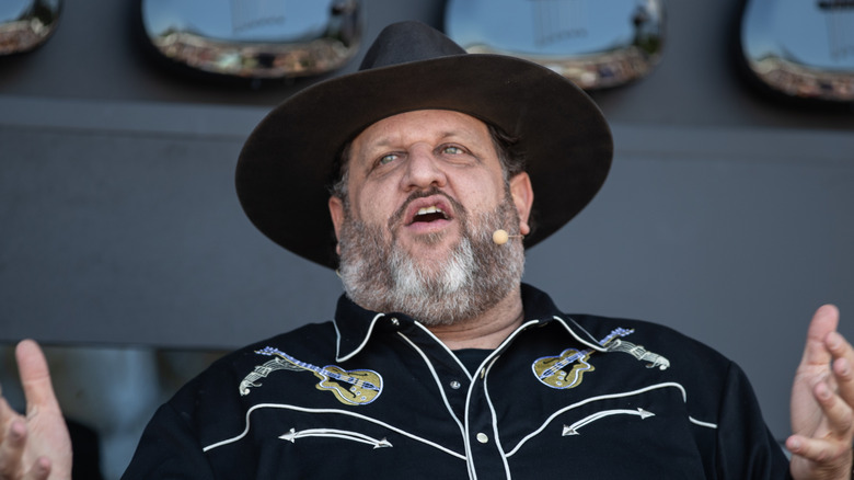 Chef Aaron May addresses an audience wearing a wide-brimmed hat and a Western shirt, with several electric guitars hung behind him