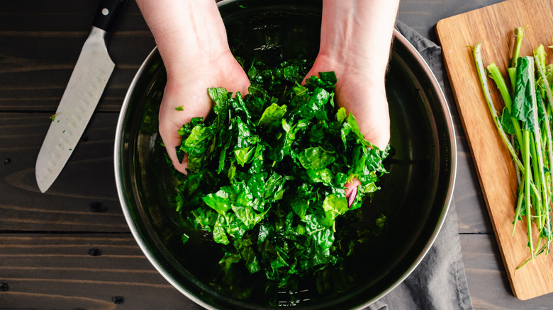 Hands massaging kale in deep bowl beside knife on wood surface.