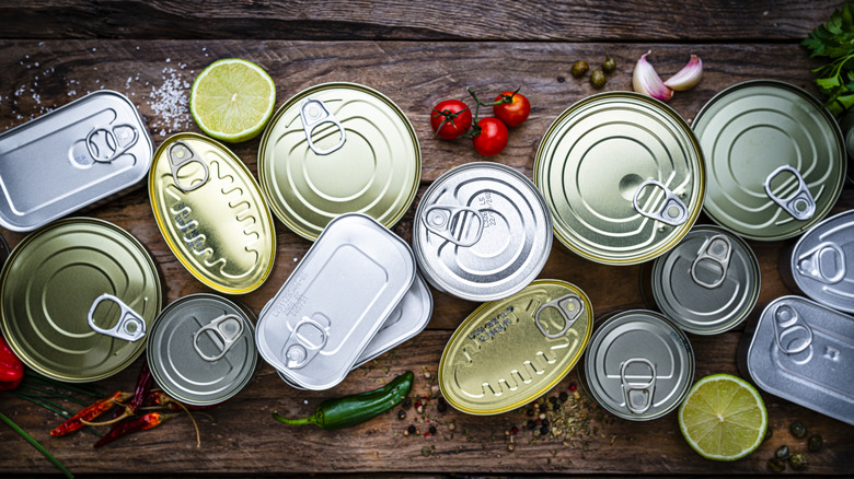 Various unlabeled cans of food on a wooden table with a few fresh ingredients