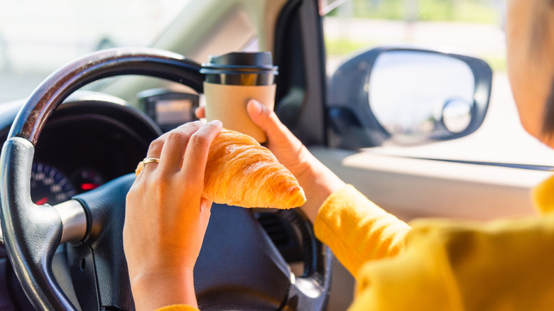 Person sitting behind the wheel holding a croissant in one hand and coffee in the other.
