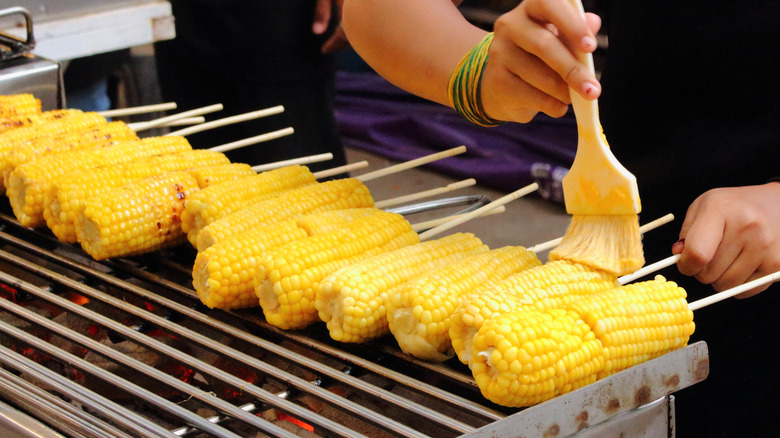 A hand uses a basting brush to coat corn cobs on a grill.