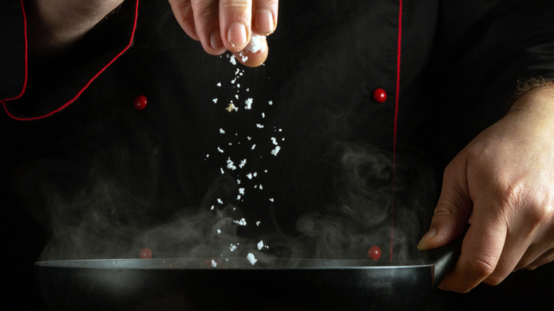 A person in a black chef's uniform sprinkles salt into a hot, steaming pan.