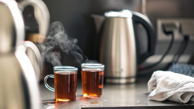 Two glass mugs of tea sit in front of a staninless steel electric kettle in a kitchen
