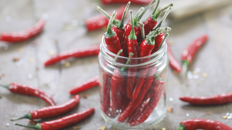 jar of hot red peppers on table scattered with more red peppers