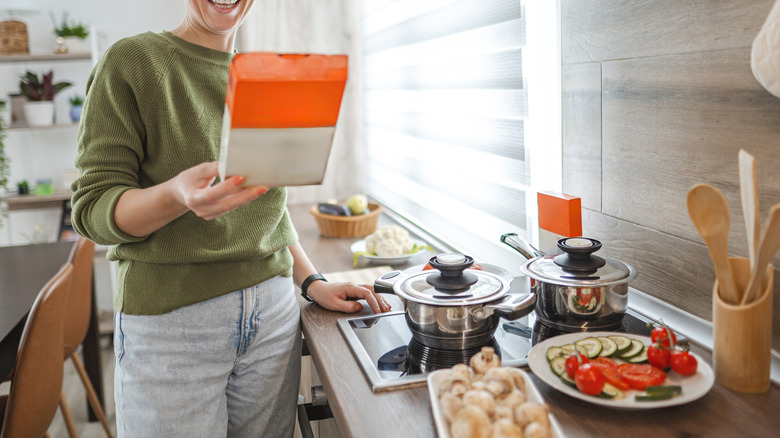 A woman wearing glasses really the instructions on the back of a box of something in her kitchen
