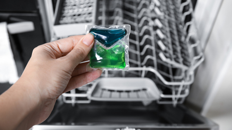 A woman's hand holding a dishwasher tablet in front of an open dishwasher