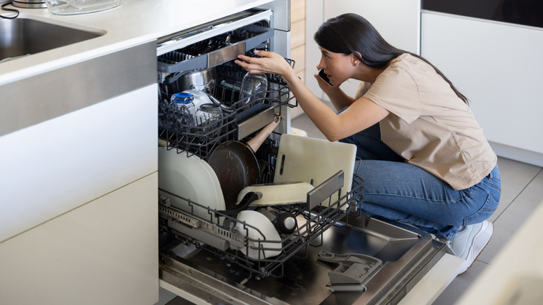 A woman in a tan top and jeans inspecting her dishwasher to check for problems.