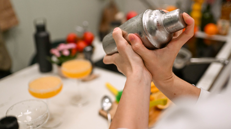 hands holding a cocktail shaker in a home kitchen with blurry cocktails in the background