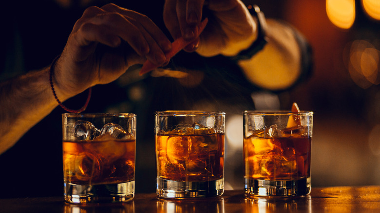 hands squeezing orange peel above three iced cocktails on a bars