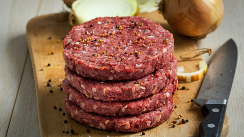 A stack of raw hamburger patties on a cutting board, seasoned with peppercorns, sitting next to some onions