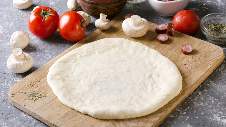 Raw pizza dough on a wooden board with tomatoes and mushrooms on the side