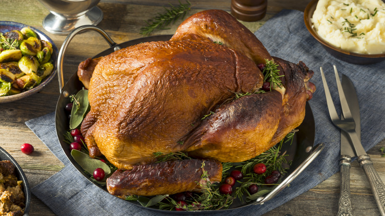 A roast turkey in a metal tray with some herbs and cranberries, on a table with mashed potatoes, brussel sprouts, and utensils